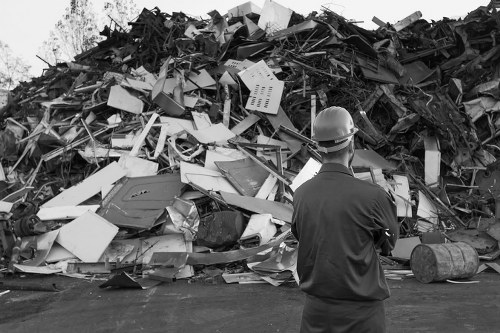A man wearing a safety helmet and dark work clothes stands with his back facing the camera, observing a large pile of mixed waste materials. The waste includes various items such as broken plastic objects, metal scraps, and debris, which are stacked in an outdoor environment, likely on a driveway or disposal area near Hoddesdon. The debris appears disorganized, with some items leaning against others, creating a chaotic landscape of discarded rubbish. The background features a clear, possibly sunny sky, providing natural light that highlights the different textures and surfaces of the waste materials. This scene reflects a typical refuse accumulation intended for removal or recycling, aligning with the rubbish removal services provided by Commercial Waste Hoddesdon, who manage commercial waste disposal in the local area.