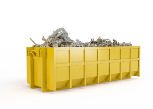 A large, rectangular skip painted in bright yellow, filled with a heap of mixed construction debris including broken concrete slabs, bricks, and fragments of plasterboard, situated against a plain white background. The debris appears loosely piled, with some pieces protruding and unevenly spread within the skip. The skip is positioned on a flat surface, with its front side facing the viewer, and features visible panels and sturdy corner supports. This scene represents typical waste removal equipment used by commercial waste services such as those provided by Commercial Waste Hoddesdon, serving clients in Hoddesdon and the surrounding areas of Hertfordshire. The natural lighting enhances the textures of the crushed materials, highlighting the rough, uneven surfaces of concrete, and the contrast between the bright yellow container and the grey debris emphasizes the clean, professional appearance of the skip for efficient rubbish collection and disposal services.