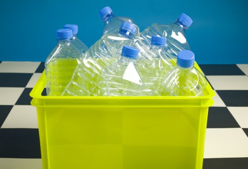A collection of clear plastic bottles with blue caps, some partially crushed, are placed inside a bright yellow plastic bin. The bottles appear to contain a transparent liquid and are arranged in a somewhat haphazard manner, with some leaning against each other. The bin is situated on a black and white checkered surface, likely a tiled floor, and the background features a solid blue wall. This image visually represents the disposal or recycling of plastic waste, which is relevant to commercial rubbish removal services in Hoddesdon, suggesting the recycling of plastics as part of waste management operations by companies such as Commercial Waste Hoddesdon.