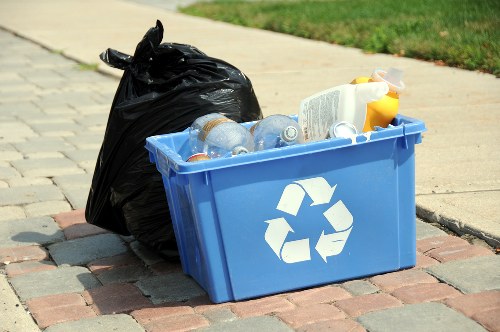 A blue plastic recycling bin with a white recycling symbol on the front, placed on a paved area beside a residential sidewalk. The bin is filled with clear and brown plastic bottles, with some caps visible, and appears to contain other mixed recyclables. Adjacent to the bin is a full black rubbish bag, tightly tied at the top, leaning against the bin. The background shows a grassy verge and a pavement, indicating an outdoor urban or suburban environment. The scene suggests waste collection or rubbish removal activity, typical of services provided by Commercial Waste Hoddesdon in the Hoddesdon area, focusing on waste management and recycling of household rubbish.