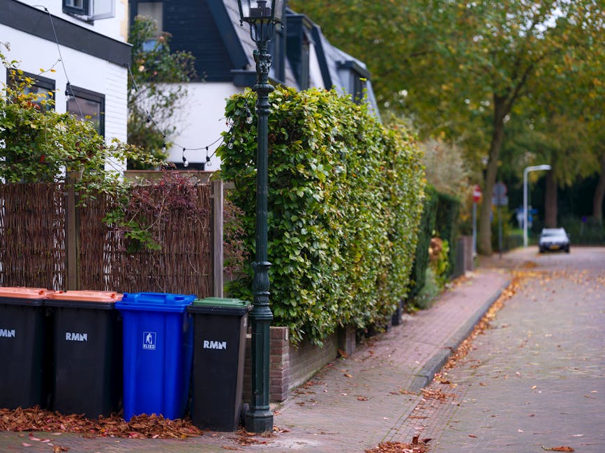 In a residential street scene, four separate wheelie bins are positioned along the edge of a sidewalk, directly beside a decorative black lamppost. The bins include two large black waste disposal cont