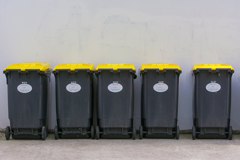 In a residential street scene, four separate wheelie bins are positioned along the edge of a sidewalk, directly beside a decorative black lamppost. The bins include two large black waste disposal cont