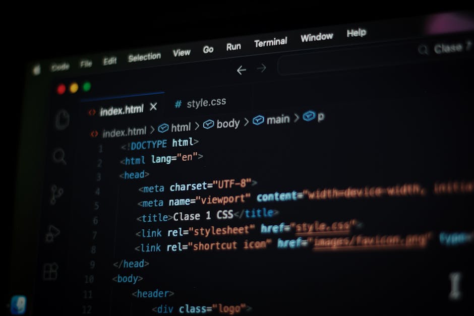 A person wearing a dark jacket and glasses is seated at a desk in an office or workplace environment, working on a computer with a large monitor displaying lines of computer code in various colors, in