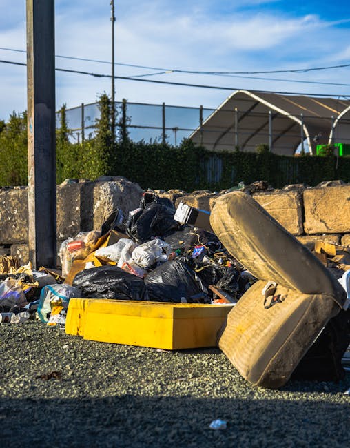 A row of large, rectangular wheelie bins lined up outdoors along a paved pathway adjacent to a brick wall. The bins are made of durable plastic and feature textured lids with ridges. The first bin in 